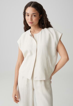 Donna con lunghi capelli ricci che indossa un top bianco senza maniche ampio e strutturato e pantaloni larghi abbinati, in piedi davanti a uno sfondo semplice.