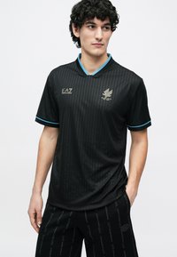 Melna sporta krekls ar plānām vertikālām svītrām, turkīza akcentiem apkaklē, zelta logotipa detaļām, īsām piedurknēm un brīvu piegriezumu.