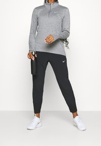 Grijze sporttop met lange mouwen en een kwart rits, gecombineerd met zwarte tapse broek. Zwarte waterfles in één hand. Witte sneakers.