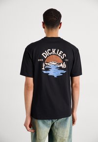 Camiseta de algodón negra con un gráfico colorido en la parte posterior que presenta montañas y agua, etiquetada con "DICKIES" en una fuente en negrita.