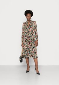 Vestido midi floral com mangas longas, decote em formato de chave, base preta, flores de várias cores e barra franzida. Combinado com saltos pretos e uma bolsa.