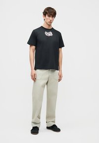 Jeune homme portant un t-shirt graphique noir, un pantalon ample beige clair et des chaussures noires, debout contre un fond clair uni.