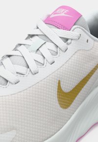Αθλητικά παπούτσια με ελαφρύ γκρι πλέγμα επάνω, χρυσό λογότυπο swoosh, ροζ λεπτομέρεια στη γλώσσα και υφασμάτινη λευκή σόλα. Τα κορδόνια είναι λευκά και επίπεδα.