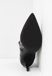 Mustat kärjellä varustetut korkokengät, jotka osoittavat ylöspäin, näyttäen sileän pohjan, jossa on merkintä koko 37, valkoista kulmatonta taustaa vasten.