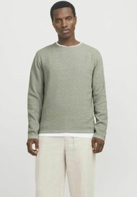 Pull en tricot vert clair avec un col rond, un motif texturé, des manches longues et un col intérieur blanc contrastant, associé à un pantalon clair.