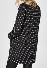 Schwarze Tunika mit weißen Polka-Dots, langen Ärmeln, oversized Design, weichem Stoff und Seitentaschen. Rückansicht gezeigt.