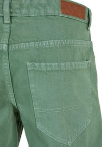 Pantalones de mezclilla verdes con etiqueta de cuero, bolsillos traseros con costura en forma de triángulo y un acabado liso y texturizado.