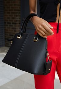 Osoba držící černou kabelku s texturou a zlatým kováním a malou připojenou taštičkou, oblečená v červených kalhotách a černém topu.