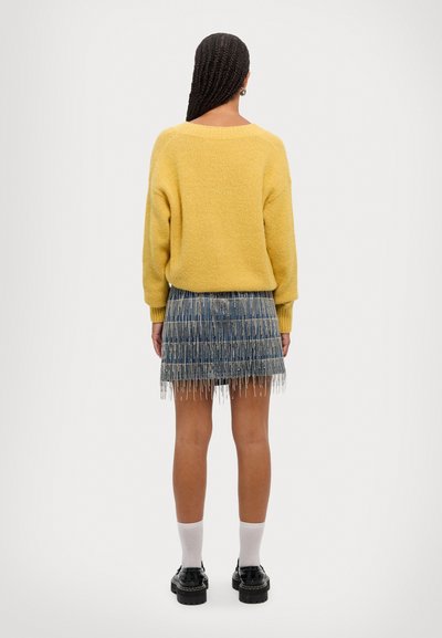 Gelber gestrickter Pullover mit weitem Ausschnitt, kombiniert mit einem blauen Rock mit strukturierten Mustern und Fransen am Saum. Schwarze Schuhe und weiße Socken.