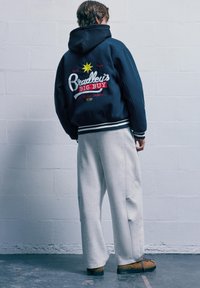 Námořnická mikina s kapucí, která má vyšívaný text, vroubkované manžety a grafickou hvězdu. Kombinováno se světle šedými širokými tepláky a tan botami.