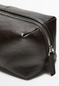 Bustina in pelle marrone scuro con texture, con cerniera argentata parzialmente chiusa, che mostra la linguetta in pelle, isolata su sfondo bianco.