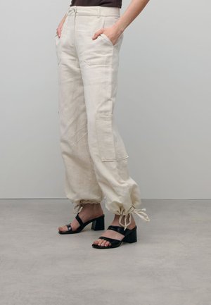 Linen, beige wide-leg pants with elastic cuffs and pockets, paired with black block-heeled sandals featuring two straps across the foot.