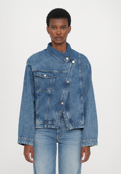 Denim jacket in medium blue with a unique asymmetric design, two front pockets, and a single button closure. Soft texture and relaxed fit.