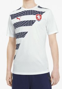 Bílý sportovní triko s námořnickými diagonálními pruhy a červeným erbem. Vyrobeno z hladkého materiálu s kulatým výstřihem a krátkými rukávy.