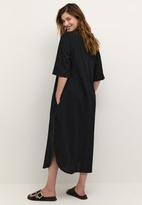 Svart lång klänning med korta ärmar, sidofickor och en slits på sidan. Slät textur, avslappnad passform och en ledig design. Bärs med platta sandaler.