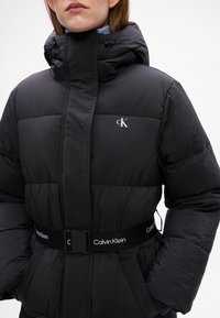 Giacca nera imbottita con colletto alto, dotata di chiusura con zip, coulisse elastiche e cintura con logo. Texture liscia, design trapuntato.