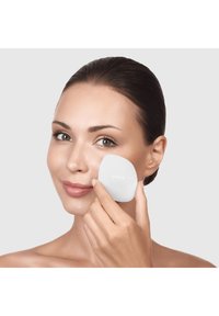 Applicateur facial en silicone blanc avec une forme douce et arrondie, tenu contre une peau lisse ; la marque est visible sur l'applicateur.