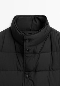 Gilet imbottito nero con colletto alto, caratterizzato da chiusure a bottoni a pressione e una texture liscia. Le sezioni trapuntate forniscono isolamento.