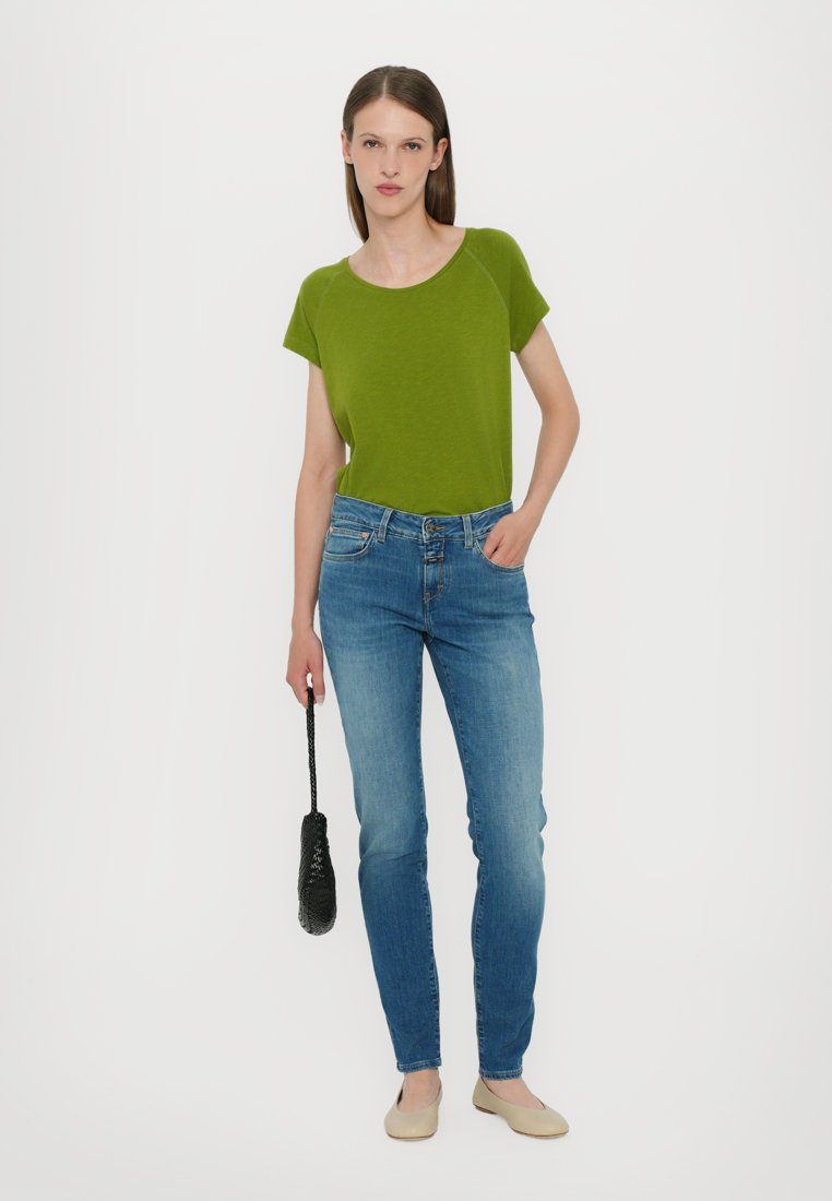 Haut vert à manches courtes, jeans bleus ajustés, chaussures claires, tenant un sac texturé noir. Design simple et décontracté avec une texture de tissu lisse.