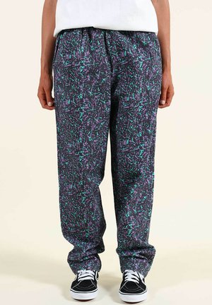 Personne portant un pantalon ample à motifs noirs, violets et turquoise avec des baskets noires et blanches, debout devant un fond uni.