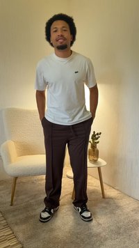 Witte T-shirt, donkerbruine broek, zwart-witte sneakers. Achtergrond met een gestructureerde muur en een lichtgekleurde, gestoffeerde stoel.