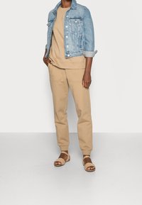 Persona che indossa una giacca di denim azzurro chiaro sopra un felpa beige e pantaloni jogger, abbinati a sandali aperti beige, in piedi davanti a un muro semplice.