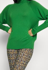 Maglione a maglia verde brillante con colletto alto, maniche raglan e vestibilità ampia. Abbinato a leggings con fantasia nei toni del blu, marrone e giallo.
