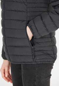 Svart puffjacka med horisontell quiltad design, blixtlåsficka på sidan och elastiska muddar. Materialet verkar slätt och isolerat.