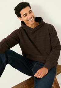 Braunes Kapuzen-Sweatshirt mit strukturiertem Muster, gerippten Bündchen und einer Fronttasche. Kombiniert mit dunkelblauen Jeans. Model sitzt auf einem Holzstuhl.