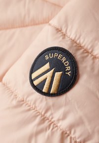 Tessuto di giacca imbottita rosa chiaro con texture trapuntata. Presenta un patch circolare blu navy con la scritta "SUPERDRY" e dettagli dorati in un design geometrico.