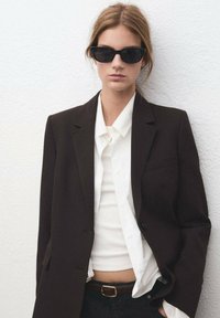 Czarny wełniany blezer na białej koszuli i prążkowanym topie na ramiączkach, zestawiony z ciemnym dżinsem. Modelka ma na sobie czarne okulary przeciwsłoneczne w kocim kształcie.