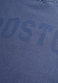 Sudadera azul con textura suave, con un estampado difuminado de "BOSTON" en la parte delantera. Cuello y puños de canalé. Diseño simple y casual.