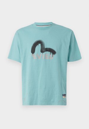 Lyseblå bomulds T-shirt med et sort og gråt spraymalet logo. Korte ærmer, rund hals og et lille mærke ved kanten.