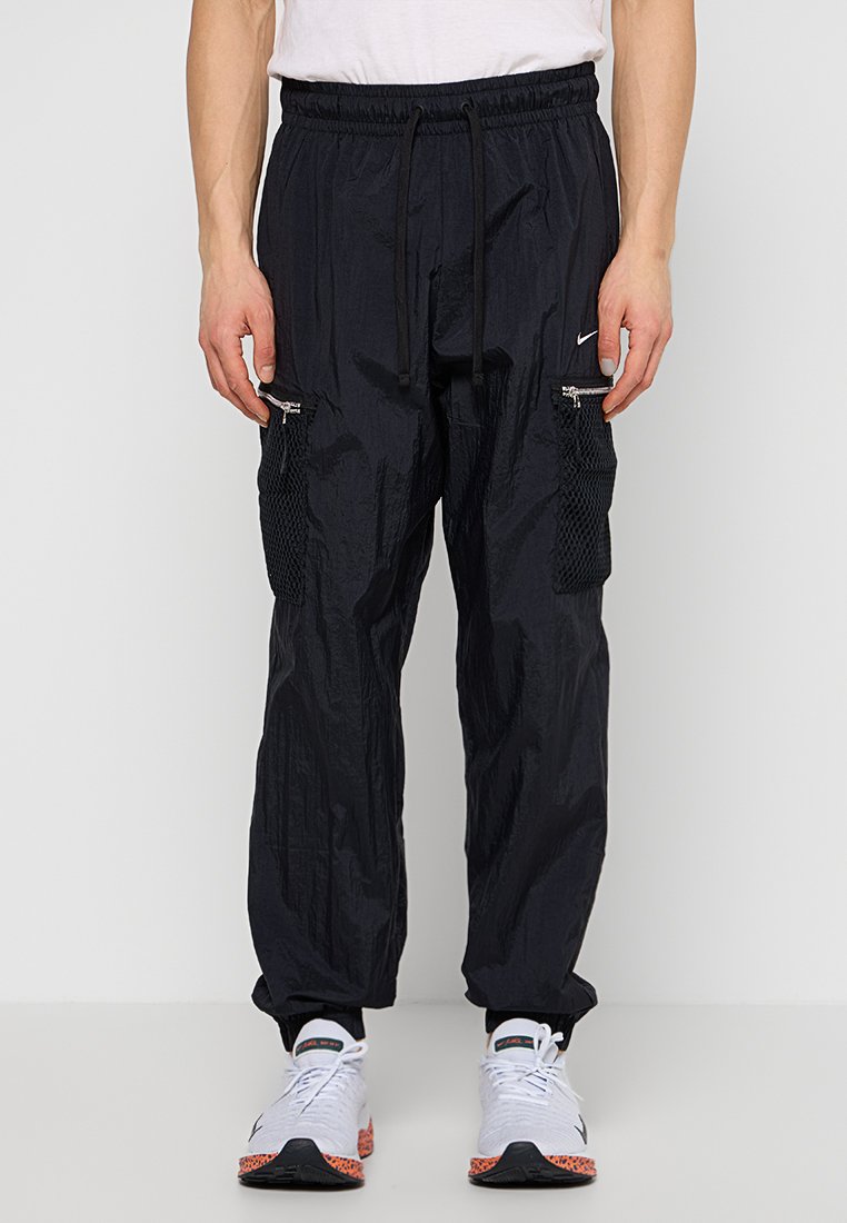 Uomo in pantaloni cargo neri con tasche in rete con zip e sneakers bianche con suole arancioni, in piedi davanti a uno sfondo semplice.