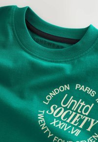 Sweatshirt vert à col rond, texture douce, présentant un imprimé graphique blanc avec du texte et des éléments de design circulaires.