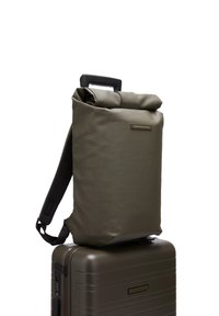 Sac à dos roulé de couleur vert olive, fabriqué dans un matériau lisse, avec une sangle unique et un accent métallique. Positionné sur une valise assortie.