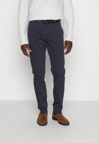 Tamsiai mėlynos medvilninės kelnės su siauru siluetu, turinčios priekinės kišenes, diržo kilpas ir derinamos su rudomis zomšinėmis bateliais.