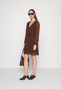 Vestido castanho com padrão, decote em V e mangas longas, combinado com tamancos pretos e uma mala preta. Os acessórios incluem óculos de sol escuros.