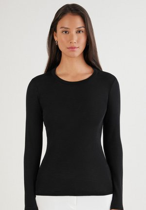 Schwarzes, langärmliges, eng anliegendes Shirt mit rundem Halsausschnitt. Glatte Textur, gerippte Bündchen und saubere Kanten. Getragen mit weißen Hosen.
