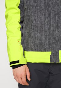 Personne portant une veste jaune et grise haute visibilité avec des poignets en Velcro et un pantalon sombre, main détendue le long du corps.