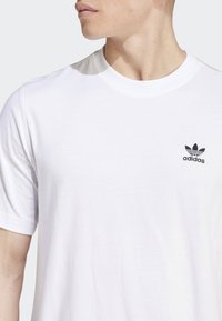 Bílý bavlněný tričko s krátkým rukávem, kulatým výstřihem a malým černým logem Adidas na levém prsu. Hladká textura, jednoduchý design.