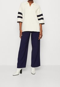 Donna che indossa una polo oversize bianca con righe nere sulle maniche, pantaloni a gamba larga blu navy e stivaletti bianchi con tacco.