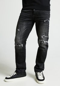 Jeans de mezclilla oscura con un acabado desgastado, que presentan rasgaduras y deshilachados en las rodillas. Combinados con zapatillas negras y blancas.