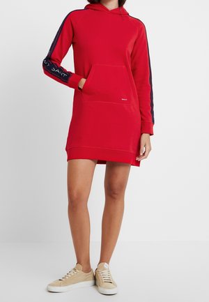 Robe sweat à capuche rouge avec une poche kangourou. Présente des bandes latérales bleu marine avec texte de logo. Fabriquée en tissu doux, design longueur genoux.