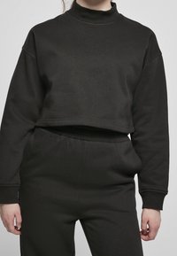 Person som bär en enkel svart kort sweatshirt och matchande högmidjade svarta byxor, stående mot en grå bakgrund.