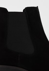 Botín de ante negro que presenta paneles elásticos en los laterales y una suela de goma negra suave. La costura es visible, realzando el diseño.