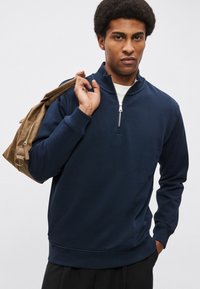 Tmavomodrá mikina na zips vyrobená z jemnej tkaniny, s vysokým golierom, dlhými rukávmi a voľným strihom. Zvýraznená strieborným zipsom.