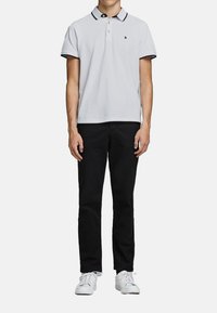 Witte poloshirt met een zwarte kraag en afwerking, voorzien van een klein logo op de borst. Zwarte broek en witte sneakers maken de outfit compleet.