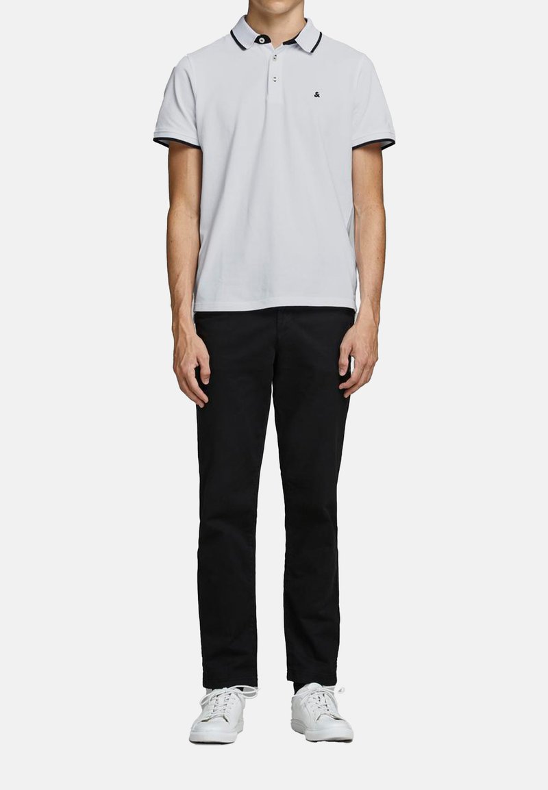 Witte poloshirt met een zwarte kraag en afwerking, voorzien van een klein logo op de borst. Zwarte broek en witte sneakers maken de outfit compleet.