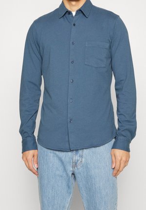 Mann trägt ein blaues Hemd mit langen Ärmeln und Brusttasche, kombiniert mit hellblauen Jeans vor einem einfarbigen Hintergrund.